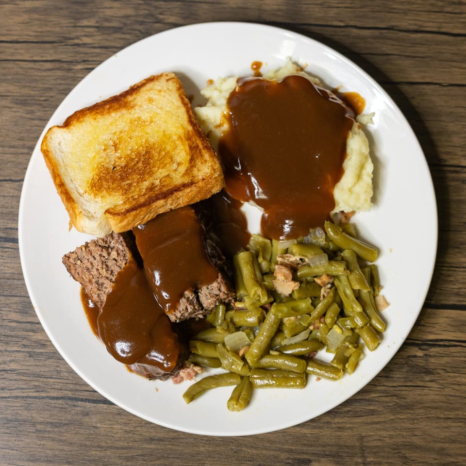Meatloaf with gravy, mashed potatoes, green beans, and toasted bread on a white plate.