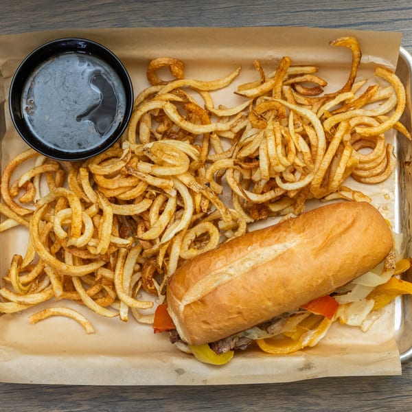 Cheesesteak sandwich with peppers and onions, curly fries, and a side of au jus on a tray.