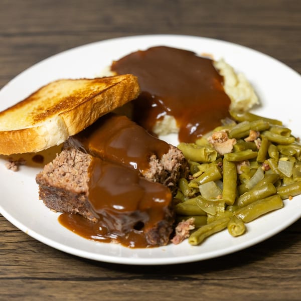 Meatloaf with gravy, mashed potatoes, green beans, and toasted bread on a white plate.