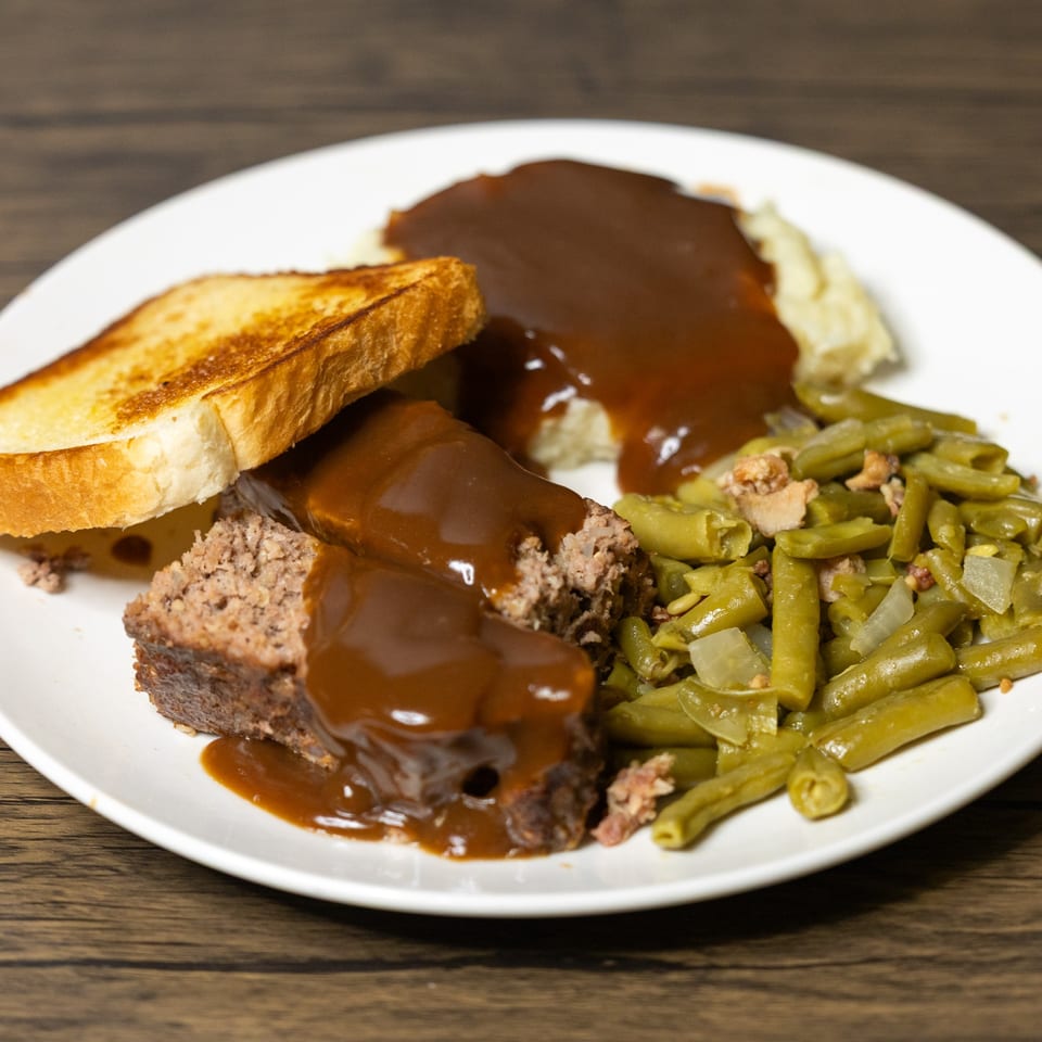Meatloaf with gravy, mashed potatoes, green beans, and toasted bread on a white plate.