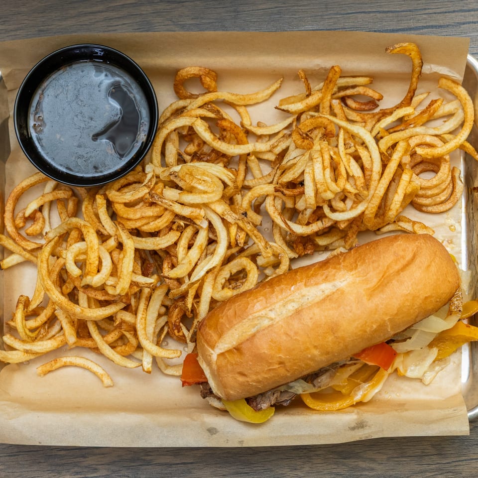 Cheesesteak sandwich with peppers and onions, curly fries, and a side of au jus on a tray.